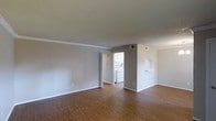 This single-floor residence offers a clean and functional layout designed for comfortable everyday living. The main living area is spacious and open, featuring warm hardwood-style flooring that extends throughout the space, creating a cohesive and inviting atmosphere. Neutral gray walls complement the bright white trim and baseboards, lending a modern yet understated aesthetic throughout.

The textured ceiling adds a classic touch, while crown molding along the upper edges of the living area provides a refined architectural detail. A pass-through opening connects the living space to the adjacent kitchen area, enhancing the sense of openness and allowing for easy interaction between rooms.

The kitchen is equipped with white appliances, including a stove and overhead microwave or range hood, set against white cabinetry. The layout is practical and efficient, making good use of the available space. A hallway visible from the main living area leads further into the residence, suggesting additional rooms beyond.

Natural light filters into the space, reinforcing the bright and airy feel of the interior. The overall ambiance is clean, neutral, and move-in ready, offering a versatile canvas well-suited for a variety of furnishing styles and personal touches.