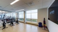 This single-floor fitness center offers a clean, modern environment designed to support a variety of workout routines. The space features a drop-panel ceiling with recessed and linear lighting that ensures consistent illumination throughout. Large windows with roller shades line the walls, inviting ample natural light and providing views of the surrounding greenery, creating an open and energizing atmosphere.

The gym is outfitted with stationary cycling bikes positioned along a mirrored wall, which enhances the sense of space and allows users to monitor their form during workouts. A dedicated free weights area includes a multi-tier dumbbell rack stocked with color-coded dumbbells in a range of weights, catering to both beginner and advanced users. Yoga and exercise mats are neatly stored nearby, making the space versatile for stretching, mat work, or group exercise.

The flooring throughout is a light-toned hardwood-style surface that is easy to maintain and comfortable underfoot. The overall design is functional and uncluttered, with neutral wall tones and a straightforward layout that maximizes usable floor space. This fitness center is well-suited for individuals seeking a practical and well-lit environment for their daily exercise needs.