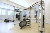 Located along Deer Trail in Milton, Georgia, this single-floor fitness facility is thoughtfully designed to support a comprehensive range of exercise activities. The space is divided into distinct functional zones, connected by an open layout that promotes visibility and flow throughout. Light hardwood-style flooring runs throughout the main exercise areas, contributing to a clean, modern aesthetic.

A prominent feature of the facility is its split-level layout, where a raised platform area—bordered by sleek stainless steel cable railings—overlooks the main workout floor below. This elevated section houses cardio equipment, including rowing machines and treadmills, and benefits from large arched and rectangular windows that frame views of the surrounding tree canopy, filling the space with natural light.

The lower workout floor accommodates strength training and functional fitness equipment, including a multi-purpose rig with integrated weight storage, a trampoline, and various free weights. Glass-paneled room dividers allow for flexible space configuration while maintaining visual openness. Recessed lighting and pendant fixtures provide consistent illumination throughout. Accessible restroom facilities are also present. The overall ambiance is bright, clean, and professionally appointed, suited for a range of individual and group fitness activities.