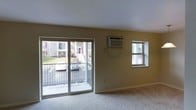 This single-floor residence offers a straightforward and functional layout designed for comfortable everyday living. The main living area is a standout feature, bathed in natural light thanks to large sliding glass doors that lead directly to a private balcony. The balcony overlooks a landscaped outdoor area, providing pleasant views of the surrounding neighborhood and greenery. A secondary window on the adjacent wall further enhances the brightness of the space, ensuring the interior feels open and airy throughout the day. The walls are finished in a warm neutral tone, complementing the carpeted flooring that runs throughout the living area, adding a layer of softness and warmth underfoot. A wall-mounted air conditioning unit is conveniently positioned for efficient climate control. The clean, simple lines of the interior reflect a no-frills aesthetic that is both practical and welcoming. The sliding doors and dual windows create a strong connection between the indoor living space and the outdoors, making natural light a defining characteristic of the overall ambiance. This space is well-suited for those seeking a comfortable, low-maintenance residence with easy outdoor access and ample natural illumination.