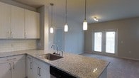 This well-appointed single-floor residence showcases a thoughtfully designed open-concept layout that seamlessly connects the kitchen and living areas. The kitchen is a standout feature, boasting granite countertops with subtle variegated tones, a stainless steel undermount sink, and a peninsula that doubles as a breakfast bar. Crisp white cabinetry with brushed nickel hardware pairs beautifully with a classic subway tile backsplash, while stainless steel appliances — including a microwave, range, and dishwasher — complete the functional yet elegant space. Three pendant lights with cylindrical shades hang above the peninsula, casting warm, focused light across the countertop. The open living area flows naturally from the kitchen, finished with plush neutral carpeting that adds warmth and comfort underfoot. Track lighting overhead provides additional illumination throughout. A set of French doors at the far end of the living space invites natural light and offers direct access to the outdoors. Neutral gray walls and clean white trim throughout create a cohesive, modern aesthetic. A hallway visible from the main living space leads to additional rooms, completing this practical and inviting floor plan.