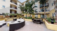 Located at 186–188 S Arroyo Pkwy in Pasadena, California, this outdoor courtyard space offers a resort-inspired retreat at the heart of a multi-story Mediterranean-style building. The courtyard is framed by warm stucco facades, arched colonnades, and wrought-iron balconies that rise across multiple levels, creating an enclosed yet open-air ambiance filled with natural light.

At the center of the courtyard, a generously sized circular wicker sectional sofa surrounds a built-in fire pit, offering a comfortable and stylish gathering area for socializing or relaxing. Adjacent to the lounge seating, a wicker dining set with a large umbrella provides a shaded space for outdoor meals and conversation.

Lush tropical landscaping is a defining feature of this courtyard, with towering queen palms, dense tropical shrubs, and richly planted garden beds lining the perimeter walls. Mulched planting areas add texture and color, contributing to a verdant, resort-like atmosphere throughout the space.

A paved brick walkway leads through the courtyard toward a gated entrance, flanked by low stucco walls and illuminated by sleek bollard lighting. The overall design blends Southern California's relaxed outdoor lifestyle with a refined architectural aesthetic, making this courtyard an inviting communal space ideal for both quiet relaxation and social gatherings.
