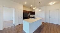 This single-floor residence is designed with an open and functional layout, connecting the main living spaces in a cohesive flow. The kitchen serves as a central highlight, anchored by a large island with a built-in sink and light countertop surface. Pendant lights hang above the island, providing focused illumination over the workspace. Dark espresso cabinetry lines the kitchen walls, offering substantial storage, while black appliances — including a refrigerator, range, and over-the-range microwave — complete the setup. Wood-style flooring runs throughout the kitchen and main living areas, lending warmth and continuity.

The bedrooms feature neutral carpeting and crisp white walls, creating a clean and adaptable environment. Windows fitted with horizontal blinds allow natural light to filter in while maintaining privacy, with views of greenery visible from at least one room.

Two bathrooms are present in the unit. One includes a tub-shower combination with tiled surround, a single-sink vanity with dark cabinetry, and wood-style flooring. The other bathroom connects directly to a walk-in closet with built-in shelving, offering convenient storage adjacent to the sleeping area.

Ceiling fans are positioned throughout the living spaces, enhancing comfort and air circulation. The overall aesthetic is consistent and neutral, characterized by white walls, dark wood accents, and clean finishes.