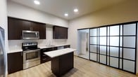 This single-floor, one-bedroom, one-bathroom residence offers a thoughtfully designed layout that maximizes both functionality and style. The kitchen serves as a central highlight, featuring dark espresso cabinetry that contrasts beautifully with light marble-toned granite countertops. A central island provides additional prep space and casual dining options, while stainless steel appliances—including a range, microwave, and dishwasher—add a polished, contemporary feel. Light-toned hardwood-style flooring runs throughout, lending warmth and visual continuity to the space. Recessed lighting illuminates the kitchen area with a clean, modern touch. A standout architectural feature is the black-framed glass partition door, which adds an industrial-chic aesthetic while allowing light to flow between rooms. The neutral wall tones create a versatile backdrop that complements a range of interior styles. The bedroom and bathroom are accessed through this striking glass door, maintaining a sense of openness while providing privacy. Overall, the space combines modern design elements with practical living features, resulting in a comfortable and visually cohesive home.