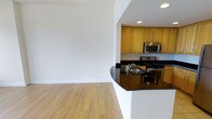 This single-floor space offers a clean, open-concept layout that seamlessly connects the living area to the kitchen, creating a sense of spaciousness and ease of flow. The living area features light hardwood flooring and crisp white walls, bathed in natural brightness that gives the space an airy, welcoming ambiance. Recessed lighting throughout ensures consistent, well-distributed illumination across the interior.

The kitchen is a standout feature, showcasing warm honey-toned wood cabinetry that provides ample storage, paired with striking dark granite countertops that add a touch of elegance and contrast. A peninsula with a curved edge extends into the open space, offering additional prep area and a natural visual divider between the kitchen and living zones. Stainless steel appliances, including a gas range and over-the-range microwave, lend a modern, functional edge to the kitchen.

The neutral color palette of white walls and light flooring throughout the space creates a versatile backdrop suitable for a variety of interior styles. The thoughtful design balances practicality with aesthetic appeal, making this a comfortable and functional single-level space.