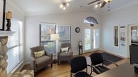 This single-story leasing office is designed to be both functional and inviting, combining professional workspaces with comfortable seating areas that create a warm atmosphere for prospective residents. The main area features vaulted ceilings accented by crown molding, wood-look flooring throughout, and a ceiling fan that enhances the open, airy feel of the space.

A standout architectural element is the rustic stone fireplace, which anchors one end of the main room and is framed by large windows fitted with horizontal blinds that allow for controlled natural light. Adjacent to the fireplace, a seating area with upholstered chairs offers a relaxed setting for consultations. The entry is highlighted by elegant French doors topped with an arched transom window, further brightening the space.

The workstation area includes a sizable desk with computer equipment and a wall-mounted flat-screen display, providing an efficient setup for leasing staff. Framed certifications and decorative mirrors adorn the walls, lending a professional yet personable touch. A secondary room off the main workspace contains a kitchenette with wood cabinetry, granite-style countertops, and black appliances, adding practical convenience to the office environment.