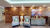 This community showcases a thoughtfully designed blend of interior and outdoor spaces that together create a resort-like atmosphere. The interior lounge area is anchored by a striking floor-to-ceiling textured wood accent wall composed of layered, varied-tone wood panels that add warmth and visual depth. Recessed lighting highlights the wall's dimension, while ambient LED cove lighting above contributes a soft glow. Two upholstered wingback chairs in light gray, accented with tropical-print throw pillows, are positioned symmetrically around a geometric side table and lamp. A coastal-themed canvas print and a credenza with decorative accessories complete the polished, inviting aesthetic.

The outdoor areas are equally impressive. A covered terrace furnished with a large sectional sofa and a round coffee table provides a comfortable gathering space beneath a pergola-style structure, framed by tall palm trees. A secondary seating nook features woven lounge chairs with cushioned seats, ideal for relaxed conversation. The centerpiece of the outdoor area is an expansive resort-style swimming pool, surrounded by a sun deck lined with chaise lounges and shade umbrellas, all set against a backdrop of multi-story buildings and lush palm landscaping.