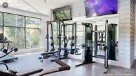 Located at Slater Crossing in Morrisville, NC, this well-appointed fitness center is organized across two distinct workout zones, offering a comprehensive and modern exercise environment. The main strength and cardio area features durable rubber flooring, large floor-to-ceiling windows that flood the space with natural light and provide lush wooded views, and shiplap-accented white walls that contribute to the clean, contemporary aesthetic. Multiple wall-mounted flat-screen TVs and a ceiling fan enhance comfort during workouts. The room houses an array of strength training equipment, including a power rack with bench press, a multi-functional cable machine, a full dumbbell rack, and various cardio machines such as treadmills and stationary bikes. A full-length mirror wall visually expands the space. The second zone features warm hardwood-style flooring, floor-to-ceiling glass partition walls, and a striking large-scale abstract floral mural that serves as a vibrant focal point. This area is equipped with a Peloton cycling bike, a rowing machine, kettlebells, medicine balls, and resistance tools, making it ideal for functional and cardio-focused training. Both zones are bright, well-ventilated, and thoughtfully designed to support a wide range of fitness activities.