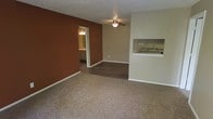 This single-story space presents an open and functional layout centered around a carpeted living area. The room is characterized by a striking terracotta-brown accent wall that adds a warm, bold contrast to the surrounding neutral beige walls. Flooring transitions from carpet in the main living zone to harder flooring in the adjacent area, delineating separate functional spaces within the open layout.

A ceiling fan with a light fixture is centrally mounted, providing both air circulation and ambient lighting. A textured popcorn ceiling runs throughout, adding a classic residential character to the space.

A pass-through window with a white countertop connects the living area to the kitchen, offering convenience and an open sightline between the two spaces. The kitchen area, visible through the pass-through, features a sink and cabinetry.

Accessible from the living area, a bathroom is partially visible through a doorway on the accent wall side, revealing a vanity with lighting above. White baseboards frame the perimeter of the room, providing a clean finish. Overall, the space offers a straightforward, practical layout with distinct design touches that create visual interest.