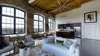 This single-floor space showcases a striking industrial-chic aesthetic, seamlessly merging raw architectural elements with warm, elegant interiors. The most defining feature is the exposed brick wall, which spans a substantial portion of the living area and anchors the space with historic character. Above, a beautifully finished wood-planked ceiling rises dramatically overhead, supported by timber beams and fitted with a large industrial ceiling fan that reinforces the loft-style atmosphere.

Oversized arched windows with black metal frames line the brick wall, flooding the interior with abundant natural light and offering open views of the surrounding landscape. The open-plan layout connects the living area directly to the kitchen, which features dark espresso cabinetry, stainless steel appliances, a subway tile backsplash, and a central island with pendant lighting overhead.

The living area is furnished with a plush white slipcovered sofa accented by richly colored throw pillows, complemented by classic wooden accent chairs and a woven area rug. Track lighting provides additional illumination, while lush greenery adds a fresh, organic touch. Overall, the space balances industrial heritage with sophisticated, livable warmth.