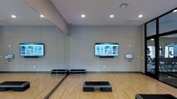 This dedicated fitness studio is designed to support a wide range of workout activities in a clean, modern environment. The space is anchored by smooth hardwood flooring that spans the entire room, providing an ideal surface for aerobics, stretching, and group exercise classes. Large wall-to-wall mirrors line one side of the studio, enhancing the sense of openness and allowing participants to monitor their form during workouts. Recessed ceiling lighting illuminates the space evenly, complemented by natural light filtering in through windows at the far end of the room. Multiple large flat-screen displays are mounted on the walls, offering access to on-demand fitness content and guided workout programs, with an interactive control panel conveniently positioned nearby. Step platforms are arranged throughout the open floor area, ready for use in structured fitness sessions. Strength and cardio equipment is visible toward the rear of the studio, extending its functionality beyond group classes. A security camera ensures a safe environment. The overall ambiance is bright, functional, and motivating, making this studio a versatile space suited for both instructor-led sessions and independent training.
