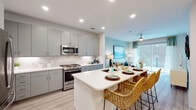 This single-floor residence is thoughtfully designed with a clean, contemporary aesthetic carried consistently throughout each space. The open-concept kitchen is a standout feature, showcasing gray flat-front cabinetry, a decorative mosaic tile backsplash, and a full suite of stainless steel appliances, including a gas range, microwave, and refrigerator. Light wood-style flooring runs throughout, brightening the space and lending a cohesive, modern feel. A generously sized white waterfall island with bar seating serves as the natural centerpiece, seamlessly connecting the kitchen to the living and dining areas.

The living area is warm and inviting, accented by a soft teal feature wall and access to an outdoor space through sliding glass doors, allowing natural light to fill the room. A ceiling fan adds both function and style overhead.

The bedroom offers a cozy retreat, distinguished by a bold dark accent wall that creates a dramatic contrast against lighter furnishings and carpeted flooring. Natural light enters through a window, and a ceiling fan ensures comfort.

Adjacent to the bedroom, the bathroom features gray cabinetry, a wide white countertop, and a large backlit mirror that enhances brightness. Light wood flooring continues here, maintaining design continuity throughout the residence.