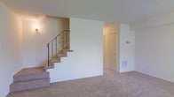 This two-floor residence offers a straightforward and functional layout with a calm, neutral aesthetic throughout. The main living area is finished with wall-to-wall carpeting in a warm beige tone, creating a soft and cohesive feel underfoot. Crisp white walls keep the space feeling clean and bright, providing a versatile backdrop for a variety of interior styles.

A carpeted staircase with a decorative wrought iron railing serves as a focal point, connecting the main level to the upper floor. The railing features elegant scrollwork detailing, adding a subtle ornamental touch to an otherwise understated space. The staircase is positioned along the side wall, allowing the main living area to remain open and unobstructed.

A doorway visible from the living area leads to an adjoining space, with a floor vent nearby indicating a central HVAC system for year-round climate control. The layout suggests a practical flow between rooms on the main level. Overall, the space presents a clean, move-in-ready environment with neutral finishes that offer flexibility for personalization across both floors.