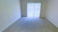 This single-story space features a bright, open room finished with wall-to-wall carpet in a neutral tone, providing a clean and versatile foundation. The room's most prominent feature is a large sliding glass door fitted with vertical blinds, which floods the interior with natural light and offers potential access to an outdoor area. The plain white walls and simple baseboard trim contribute to a minimalist aesthetic, keeping the space feeling open and adaptable. The overall ambiance is clean and understated, offering a blank canvas well-suited for a variety of furnishing and design arrangements.