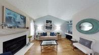 This single-floor space showcases a welcoming living room anchored by a striking vaulted ceiling that adds a strong sense of height and openness. The ceiling's angular peak draws the eye upward, creating an airy, expansive atmosphere despite the room's intimate scale. Light blue accent walls complement the crisp white upper walls and ceiling, lending a calm, coastal-inspired ambiance throughout the space.

The living room features warm hardwood flooring that runs throughout, providing a natural contrast to the cool color palette. A classic white fireplace with a black hearth serves as a focal point along one wall, flanked by a window dressed with bold patterned drapes that introduce additional visual interest. Built-in shelving on the opposite wall offers practical display space.

Furnishings include a neutral beige sofa accented with navy blue patterned throw pillows, paired with mirrored side tables and a sleek glass-and-metal coffee table that reflect light and enhance the room's brightness. Wall-mounted sconces and floor lamps provide layered lighting. Abstract artwork in coordinating blue tones ties the design together, creating a cohesive, well-styled interior with a contemporary yet comfortable character.