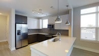 Located at the North Carolina Music Factory in Charlotte's vibrant NoDa-adjacent district, this single-floor residence offers a sleek, modern living experience defined by clean lines and contemporary design. The open-concept kitchen serves as a striking centerpiece, showcasing high-gloss white upper cabinetry contrasted by rich dark lower cabinets and complemented by a gray tile backsplash. A generous island with a bright white quartz countertop provides ample workspace and casual seating, anchored by an undermount sink with a polished chrome arc faucet. Pendant lighting in a brushed industrial style hangs above the island, adding both function and visual character. Stainless steel appliances, including a full-size refrigerator, built-in microwave, and range, deliver a refined, modern aesthetic throughout. Track lighting along the ceiling highlights the kitchen's open flow into the living areas. Light hardwood-style flooring runs throughout, lending warmth to the otherwise crisp, modern palette. Natural light filters in through a window above the kitchen, keeping the space feeling bright and airy. The overall ambiance is polished and contemporary, ideal for those seeking a stylish urban home in one of Charlotte's most energetic and culturally rich neighborhoods.