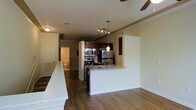 This single-floor space showcases an open-concept layout that integrates the kitchen and living areas into one cohesive environment. The kitchen is well-appointed with stainless steel appliances, including a refrigerator, range, and built-in microwave, all complemented by rich dark wood cabinetry. A granite-topped peninsula with an integrated sink serves as both a functional workspace and a casual dining or entertaining bar, separating the kitchen from the main living area. Pendant lighting hangs above the peninsula, adding focused illumination, while a ceiling fan with additional track lighting overhead provides both air circulation and ambient light throughout the space. The warm-toned hardwood flooring runs consistently across the open area, contributing to a unified and inviting atmosphere. Crown molding details along the upper walls add a refined architectural touch. A hallway visible in the background leads to additional rooms, including what appears to be a bathroom, suggesting a practical and efficient floor plan. The neutral wall tones throughout keep the space feeling bright and versatile.