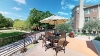 This inviting outdoor patio space offers a comfortable and functional setting for outdoor leisure and social gatherings. The patio is finished with a warm-toned concrete surface and is furnished with round tables paired with sling-back chairs, each shaded by large striped market umbrellas that provide ample protection from the sun. Multiple seating groupings are arranged across the space, accommodating both intimate conversations and larger gatherings.

The surrounding landscape is thoughtfully maintained, featuring decorative rock beds, seasonal flowering plants in vibrant pots, and a variety of mature trees that provide a lush green backdrop. A low metal railing defines the perimeter of the patio, offering an open yet defined boundary that transitions naturally into the surrounding grounds.

Adjacent to the patio is a multi-story building constructed with classic brick and neutral-toned siding, lending an established and refined architectural character to the setting. The outdoor area benefits from abundant natural light under open skies, making it a bright and cheerful environment throughout the day. The combination of structured hardscaping, verdant plantings, and well-chosen outdoor furnishings creates a welcoming atmosphere that encourages outdoor enjoyment in a well-kept, community-oriented setting.