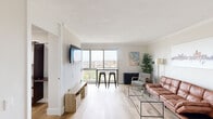 This well-appointed single-floor unit offers a bright and contemporary living experience anchored by an open-concept main living area. Light hardwood flooring runs throughout, creating a warm, cohesive aesthetic that ties each room together seamlessly.

The spacious living room is the centerpiece of the unit, featuring large floor-to-ceiling windows that frame sweeping city views and flood the space with natural light. A wall-mounted flat-screen TV, a sleek media console, and a leather sofa arranged around a glass coffee table make this an inviting area for relaxing or entertaining. A bar-height table with industrial-style stools sits directly in front of the windows, serving as an ideal dining or casual workspace with a view.

One corner of the living area transitions into a functional home office zone, complete with wall-mounted shelving and a dedicated desk and ergonomic chair — a thoughtful addition for remote work. Crown molding lends a polished, refined touch throughout.

The bedroom is a calm, restful retreat with its own large window offering more cityscape views. A natural wood platform bed, soft neutral tones, and clean crown molding detailing contribute to the room's serene atmosphere.

The kitchen features slate-grey cabinetry, white countertops, stainless steel sink, and black appliances, offering a compact yet fully equipped cooking space with crown molding accents that maintain the unit's cohesive design language.
