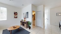This two-story home offers a clean, contemporary aesthetic defined by crisp white walls, light-toned tile flooring, and an abundance of natural light throughout. The main living area sets a welcoming tone with its open layout, featuring smooth tile floors that extend seamlessly from room to room. Soaring walls and high ceilings amplify the sense of space, creating an airy, uncluttered atmosphere. Decorative touches such as bold wall art add personality without overwhelming the neutral palette. A louvered bi-fold closet door provides functional storage while maintaining the home's understated style. Adjacent to the living area, a passageway leads to a well-lit bathroom or utility space, finished with bright cabinetry and track lighting overhead. A glimpse toward the exterior hints at an outdoor area accessible through large windows or glass doors, inviting natural light deep into the interior. The overall design balances simplicity with comfort, making thoughtful use of each space across both floors. Light, openness, and a cohesive neutral color scheme give this home a versatile and inviting character suited to a variety of lifestyles.