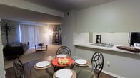 This single-story space showcases an open-concept layout that seamlessly connects the dining area, living room, and kitchen. The dining area features hard flooring and is furnished with a round glass table paired with decorative metal chairs, positioned adjacent to a pass-through counter that connects to the kitchen. This countertop bar provides a convenient transition between the two spaces, adding both functionality and openness to the floor plan.

The living area extends beyond the dining space and is finished with wall-to-wall carpeting, lending a warm, comfortable feel. Vertical blinds adorn a large sliding glass door, which allows natural light to filter into the space while offering access to the outdoors. A shelving unit in the corner of the living room adds a decorative storage element.

Throughout the space, neutral-toned walls contribute to a calm, cohesive atmosphere. The textured ceiling adds subtle dimension overhead. The open flow between rooms creates a sense of spaciousness, making the layout practical for both everyday living and entertaining. Overall, the space presents a functional and well-connected interior design.