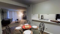 This single-story space showcases an open-concept layout that seamlessly connects the dining area, living room, and kitchen. The dining area features hard flooring and is furnished with a round glass table paired with decorative metal chairs, positioned adjacent to a pass-through counter that connects to the kitchen. This countertop bar provides a convenient transition between the two spaces, adding both functionality and openness to the floor plan.

The living area extends beyond the dining space and is finished with wall-to-wall carpeting, lending a warm, comfortable feel. Vertical blinds adorn a large sliding glass door, which allows natural light to filter into the space while offering access to the outdoors. A shelving unit in the corner of the living room adds a decorative storage element.

Throughout the space, neutral-toned walls contribute to a calm, cohesive atmosphere. The textured ceiling adds subtle dimension overhead. The open flow between rooms creates a sense of spaciousness, making the layout practical for both everyday living and entertaining. Overall, the space presents a functional and well-connected interior design.
