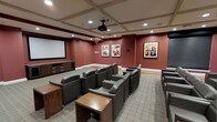 This private screening room is thoughtfully designed to deliver a premium cinematic experience. The space is anchored by a large projection screen mounted on a deep red accent wall, paired with a ceiling-mounted projector that ensures crisp, high-quality viewing. Rich, dark leather recliner-style chairs are arranged in a tiered row layout, each accompanied by wooden side tables for added convenience. The warm, neutral-toned carpet and bold burgundy walls create an intimate and immersive atmosphere reminiscent of a boutique movie theater. The ceiling features elegant coffered detailing with recessed lighting, adding architectural character and a refined sense of depth to the room. A wood media console positioned along the side wall houses additional audio-visual equipment, ensuring seamless presentation capabilities. The walls are adorned with framed pop-art style portraits, adding a stylish, curated touch to the décor. The overall ambiance strikes a balance between comfort and sophistication, making this screening room an ideal venue for private film screenings, presentations, or entertainment gatherings.