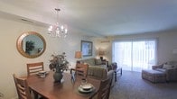This two-floor residence features a welcoming open-concept main living area that seamlessly connects the dining and living spaces. The dining area is anchored by a classic chandelier with candelabra-style lighting, casting a warm glow over a wooden dining table with cushioned chairs. Decorative touches, including a floral centerpiece and framed tableware, add a homey character to the space.

The adjoining living room is carpeted and comfortably appointed with a neutral-toned sofa, accent chairs, and table lamps that contribute to the room's warm ambiance. A large sliding glass door along the far wall allows generous natural light to fill the space and provides access to an outdoor area, enhancing the sense of openness. Vertical blinds offer privacy and light control as needed.

The walls are painted in a soft, neutral tone, complementing the warm furnishings throughout. A framed artwork piece above the sofa adds a subtle decorative element. The overall atmosphere of the main living area is cozy and functional, designed to support both everyday living and casual entertaining across its two floors.
