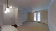 This two-story home combines traditional design elements with comfortable, functional living spaces throughout. The interior is characterized by neutral, warm-toned walls that create a welcoming and versatile backdrop for any décor style. Crown molding runs along the ceilings, adding a refined architectural detail that elevates the overall aesthetic of the space. The flooring transitions thoughtfully between plush carpeting in the main living areas and hardwood in connecting hallways or entryways, offering both comfort and durability. A ceiling fan with a built-in light fixture serves as a practical focal point in the main living area, promoting air circulation and illumination. French doors with slatted blinds provide access to an outdoor area while allowing natural light to filter into the room, contributing to a bright and airy ambiance. White panel doors and crisp white baseboards contrast cleanly against the neutral walls, reinforcing the home's classic, cohesive design language. The layout across two floors allows for a clear separation between living and private spaces, making the home well-suited for both everyday living and entertaining.