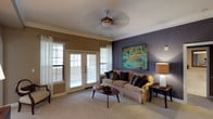 This single-story Dallas property offers a thoughtfully designed living space that blends comfort with contemporary style. The living room serves as the centerpiece of the home, immediately drawing attention with a bold purple accent wall adorned with a large, vibrant abstract painting in teal and gold tones. Crown molding runs along the ceiling perimeter, adding an elegant architectural detail that complements the neutral white ceiling above. A ceiling fan with a brushed metal finish provides both function and visual interest overhead. Natural light flows generously into the room through multiple windows fitted with horizontal blinds, as well as a set of classic French doors that open to an outdoor area, enhancing the sense of openness. The soft, neutral carpeting underfoot contributes to a warm and inviting atmosphere. The room is furnished with a plush, tan upholstered sofa accented with dark decorative pillows, paired with a mid-century style oval coffee table. A tall table lamp with an amber shade adds warm ambient lighting in the corner. The overall ambiance strikes a balance between relaxed and refined, making it a comfortable and welcoming living environment.