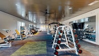 This 24-hour fitness center in Vestavia Hills, Alabama offers a generously proportioned, single-floor layout designed to accommodate a full range of workout needs. The space features a clean, modern aesthetic with crisp white walls, a distinctive wood-paneled ceiling accented by ceiling fans and track lighting, and vibrant multi-toned carpet tiles that visually define different workout zones throughout the floor.

The cardio area is lined with large windows that flood the space with natural light and provide exterior views, housing a row of treadmills, stair climbers, and elliptical machines. The strength and resistance training section is well-appointed with Matrix-brand selectorized machines, a dumbbell rack with a full weight range, adjustable benches, and a dedicated power rack. A functional training zone rounds out the facility, stocked with kettlebells, medicine balls, resistance bands, plyo boxes, and stability balls stored on organized racks.

A large wall-mounted television provides entertainment throughout the main training area, while floor-to-ceiling glass-paneled interior doors create an open, connected feel between sections of the facility. A storage closet supports operational needs. The overall ambiance is bright, clean, and motivating, making this fitness center a comprehensive and accessible resource for members around the clock.