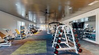 This 24-hour fitness center in Vestavia Hills, Alabama offers a generously proportioned, single-floor layout designed to accommodate a full range of workout needs. The space features a clean, modern aesthetic with crisp white walls, a distinctive wood-paneled ceiling accented by ceiling fans and track lighting, and vibrant multi-toned carpet tiles that visually define different workout zones throughout the floor.

The cardio area is lined with large windows that flood the space with natural light and provide exterior views, housing a row of treadmills, stair climbers, and elliptical machines. The strength and resistance training section is well-appointed with Matrix-brand selectorized machines, a dumbbell rack with a full weight range, adjustable benches, and a dedicated power rack. A functional training zone rounds out the facility, stocked with kettlebells, medicine balls, resistance bands, plyo boxes, and stability balls stored on organized racks.

A large wall-mounted television provides entertainment throughout the main training area, while floor-to-ceiling glass-paneled interior doors create an open, connected feel between sections of the facility. A storage closet supports operational needs. The overall ambiance is bright, clean, and motivating, making this fitness center a comprehensive and accessible resource for members around the clock.