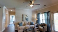 This single-floor space exudes a relaxed coastal charm, blending comfort and style in a thoughtfully curated environment. The living area serves as the heart of the space, featuring a plush cream sofa adorned with teal and patterned accent pillows that echo a seaside aesthetic. A dark wood coffee table anchors the seating arrangement, while a woven rattan armchair adds natural texture and warmth. Crown molding frames the room's perimeter, lending an elegant architectural touch, and a ceiling fan with a built-in light fixture provides both function and a classic finishing detail. Soft, neutral walls create a bright and airy backdrop, complemented by natural light filtering through windows dressed in rich teal drapes. Coastal-themed artwork — including a striking heron painting — adds personality and reinforces the overall design narrative. Warm-toned table lamps contribute to the inviting ambiance during evening hours. The dark hardwood flooring grounds the space, providing a rich contrast to the lighter furnishings and walls. Overall, this space offers a serene, beach-inspired retreat that balances casual comfort with refined design details.