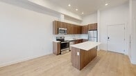Located at 1925 W North Temple in Salt Lake City, UT, this modern one-bedroom, one-bathroom unit offers a well-designed open-concept layout across a single floor. The combined living, dining, and kitchen area serves as the heart of the home, featuring light hardwood-style flooring, recessed lighting, and crisp white walls that create a bright, clean aesthetic throughout. The kitchen is outfitted with warm walnut-toned cabinetry, white quartz countertops, a white subway tile backsplash, and a full suite of stainless steel appliances including a range, microwave, dishwasher, and refrigerator. A central kitchen island provides additional prep space and a natural gathering point for the open living area. Large windows allow natural light to flow into the living space, with views of the surrounding Salt Lake City neighborhood. The bedroom is a comfortable, light-filled room with a broad window fitted with blinds and the same hardwood-style flooring found throughout. Two closets provide ample storage. The bathroom features a white tiled tub-shower combo, a walnut vanity with quartz countertop, and a large mirror, maintaining the unit's clean, modern design language. Additional conveniences include a dedicated laundry area, a hallway, and a private balcony accessible from the main living space.