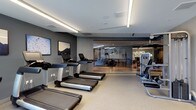 This fitness facility presents a thoughtfully designed workout space with a clean, contemporary aesthetic. The main exercise area features a row of treadmills and cardio machines along one wall, complemented by strength training equipment along the opposite side, providing a balanced layout for a full-body workout experience. The space is finished with light-toned flooring suited for gym use, while the walls are painted in a cool, neutral grey that contributes to a focused and modern atmosphere. Recessed ceiling lighting and linear LED fixtures illuminate the space evenly, ensuring a bright and functional environment. Framed artwork on the walls adds a subtle decorative touch, softening the utilitarian nature of the space. A glass partition wall separates the main gym floor from an adjacent area, which appears to serve as a lounge or multipurpose room, featuring warmer wood-toned flooring and casual seating. This open visual connection between the two zones enhances the sense of spaciousness throughout. Overall, the facility is well-organized, clean, and designed to support both cardio and strength training needs.