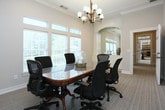 This single-floor meeting space is a thoughtfully designed conference room that combines professional functionality with refined aesthetic details. At its center sits a polished hardwood conference table with a rich, reflective finish, surrounded by ergonomic mesh and leather office chairs that comfortably accommodate multiple attendees.

The room is bathed in natural light, courtesy of a generous wall of windows fitted with adjustable horizontal blinds. Transom windows above the main panes allow additional light to filter in while offering views of the surrounding greenery outdoors. Crown molding lines the ceiling throughout, lending an air of architectural sophistication to the space.

A statement brass chandelier with frosted glass shades serves as the focal point overhead, providing warm ambient illumination that complements the natural light. On the opposite wall, a triptych of framed feather artwork adds a curated, contemporary touch against the soft neutral-toned walls.

A graceful arched passageway with decorative columns connects the conference room to an adjoining built-in display niche, hinting at a broader, well-considered floor plan. Neutral carpeting grounds the space and contributes to its professional atmosphere. The combination of classic architectural elements, plentiful natural light, and carefully chosen décor makes this a highly functional and visually inviting space for meetings and collaborative work.