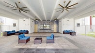 Located at 25850 SW 140th Ct in Homestead, Florida, this single-level communal amenity space offers a welcoming and well-appointed environment designed for relaxation and social gathering. The space is defined by its expansive covered pavilion structure, supported by clean white columns and exposed ceiling beams that lend an open, airy architectural character throughout.

Large-format stone-look tile flooring spans the entire area, creating a seamless and polished surface that transitions smoothly between indoor and outdoor zones. Multiple seating arrangements featuring wicker furniture with deep blue cushions are thoughtfully distributed across the space, offering comfortable gathering spots for residents and guests.

A striking blue and gold marbled accent wall serves as a bold focal point in the main interior section, complemented by oversized ceiling fans that enhance air circulation. The covered outdoor portion opens directly to views of a resort-style swimming pool, lush palm trees, and the surrounding residential buildings, bringing a distinctly South Florida atmosphere to the setting.

White metal railings define the perimeter of the open-air sections, providing a sense of enclosure while maintaining unobstructed sightlines to the pool area and tropical landscaping beyond. The overall ambiance is bright, clean, and inviting, reflecting a modern community design well-suited to Florida's warm climate.