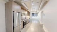 This thoughtfully designed one-bedroom, one-bathroom unit at 267 Main St in New Britain, CT offers a contemporary living experience with a clean, industrial-modern aesthetic. The open-concept living and kitchen area features polished concrete floors, exposed ductwork along a white paneled ceiling, and track lighting that illuminates the space efficiently. The kitchen is outfitted with light wood cabinetry, sleek gray countertops, a tile backsplash, and a full suite of stainless steel appliances including a refrigerator, electric range, and over-the-range microwave. A window at the end of the main living area draws in natural light and provides street views. The bedroom offers warm light hardwood flooring, crisp white walls, and a large window that further brightens the room. A dedicated closet provides practical storage. The bathroom is finished with large-format stone-look tile surrounding a soaking tub and shower combination, complemented by matte black fixtures, a wall-mounted toilet, and a vanity with a light wood base. Connecting hallways tie the layout together cohesively, making this a well-appointed and stylish urban residence.