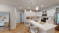 Located at 7111 Rock Fish Ln in Leland, NC, this single-floor home offers a thoughtfully designed layout with clean, contemporary finishes throughout. The kitchen is a standout feature, boasting crisp white shaker cabinetry with glass-front upper panels, quartz countertops, stainless steel appliances, a deep undermount sink, and a generous island with pendant lighting and bar seating — all set atop warm light hardwood flooring. The living and dining area is bright and welcoming, with recessed lighting, a ceiling fan, and French doors that open to an outdoor deck, flooding the space with natural light. The bedrooms offer comfortable retreats; one features a bold navy accent wall paired with natural light from large windows, while another includes a ceiling fan and access to a spacious walk-in closet with wire shelving. The bathrooms are finished with white cabinetry, quartz vanity tops, and hardwood-style flooring. The primary bath includes a large glass-enclosed walk-in shower, while the secondary bath provides a full vanity and natural light via a window. A roomy walk-in closet adds practical storage. The home's consistent use of neutral tones, quality finishes, and coastal-inspired accents creates a cohesive and inviting atmosphere throughout.