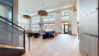 Located in Baltimore, MD, this generously sized bar and billiards room is a masterfully designed amenity space that blends industrial character with refined comfort. The space is anchored by dramatic double-height ceilings lined with oversized multi-pane windows that flood the interior with abundant natural light and offer views of the surrounding cityscape. Wide-plank hardwood flooring flows throughout, unifying the various zones within the space.

The main lounge area features multiple seating arrangements with plush sofas, leather club chairs, and layered area rugs, all centered beneath striking large-format pendant light fixtures with warm metallic finishes. A dedicated bar area showcases a substantial counter with stone tops, industrial-style cabinetry, and soaring floor-to-ceiling windows framed by overhead garage-style doors that enhance the open, airy feel.

The billiards zone offers a full-size pool table set on a rich herringbone wood floor inlay, flanked by built-in banquette seating and mirrored wall panels under a warm wood-paneled ceiling. A private dining room with a wood-beamed ceiling, large glass-top conference table, and built-in dark cabinetry provides a more intimate setting. A metal staircase with perforated railings connects to an upper mezzanine level, adding architectural depth to this exceptional communal space.