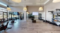 This contemporary fitness studio is thoughtfully designed to support a wide range of workout disciplines within a single, open-plan space. The studio is unified by light-toned hardwood flooring and high ceilings that lend an airy, expansive feel throughout.

The cycling area is positioned along a floor-to-ceiling glass curtain wall, flooding the space with natural light and offering views of the surrounding greenery — creating an energizing environment for spin classes. Stylish oversized pendant lamps hang throughout, adding a refined aesthetic touch that sets this studio apart from a typical gym setting.

A large open training floor occupies the central zone, bordered by full-length wall mirrors that enhance the sense of space and allow for form monitoring during workouts. A wall-mounted flat-screen display and projection screen support instructor-led classes. A ballet barre runs along the mirrored wall, making the space well-suited for barre, yoga, or stretching sessions.

Along one wall, a robust functional training rack holds an organized array of kettlebells, resistance tools, medicine balls, and stability balls, providing ample equipment for strength and conditioning workouts. Glass-paneled double doors connect the studio to an adjacent weight room, offering seamless access to additional fitness amenities. The overall ambiance is clean, modern, and inviting.