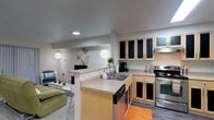 This single-floor unit is thoughtfully designed to maximize functionality within a compact footprint. The open-concept layout connects the kitchen and living area seamlessly, with a peninsula counter serving as a natural divider between the two spaces. The kitchen is well-equipped with stainless steel appliances, including a range, refrigerator, and microwave, complemented by light wood cabinetry with dark-paneled upper cabinet doors and wood-look flooring throughout. A double-basin sink with a faucet is integrated into the peninsula, adding convenience to the kitchen workspace.

The living area features soft carpeting and is furnished with a green sofa, a glass coffee table, and wall-mounted lighting fixtures that provide a warm ambiance. A built-in desk and shelving unit along one wall create a dedicated study or workspace, making the space well-suited for both relaxing and working.

The sleeping area is carpeted and includes a spacious closet with shelving, vertical blinds on the window, and room for seating. The bathroom offers a wide vanity with a large mirror, wood-look flooring, and bright overhead lighting. Recessed ceiling lights throughout the unit ensure consistent illumination across all areas.