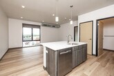 This contemporary 1-bedroom, 1-bathroom unit offers a well-designed open-concept layout that efficiently combines the kitchen and living area into a single, cohesive space. Light hardwood flooring runs throughout, creating a warm and consistent aesthetic across every room. The kitchen is a functional centerpiece, anchored by a large island with a white quartz countertop, an integrated sink, and a dishwasher. Wood-toned cabinetry complements a full suite of stainless steel appliances, including a refrigerator, electric range, and over-the-range microwave. Pendant lighting above the island adds a modern, finished touch. Exposed cylindrical ductwork runs through the living area and bedroom, lending an urban, industrial character to the space. Large black-framed windows in both the living area and bedroom invite abundant natural light and frame elevated views of the surrounding cityscape. The bedroom is a well-proportioned private retreat with its own window and access to a dedicated closet. The bathroom features wood-toned cabinetry with a white countertop vanity, hardwood flooring, and clean white walls, maintaining the unit's modern design language throughout.