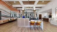 Located at 8700 Stacy Rd in McKinney, TX, this 1,710-square-foot single-floor community or leasing center is a showcase of contemporary design. The open-concept layout flows seamlessly across light-toned hardwood flooring, connecting distinct functional zones with a warm, cohesive aesthetic. The grand reception area commands attention with soaring coffered ceilings accented by slatted wood inlays, structural columns, and a dramatic chandelier. A bold, floor-to-ceiling mural wall featuring richly layered earth-tone striations serves as a stunning focal point behind the sleek white reception desk. A dedicated Brew Bar with bar-height seating and open shelving provides a stylish and functional hospitality feature. The lounge area is appointed with cognac leather sofas, quilted accent chairs, and a substantial round wood coffee table, all bathed in abundant natural light from oversized windows overlooking lush, tree-lined outdoor views. A separate sitting nook with a dining-height table and pendant lighting offers additional gathering space. An arched doorway leads to a private office or workspace, adding architectural character. Curated artwork throughout completes the refined, inviting atmosphere of this well-appointed space.