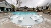 Located at 1235 Ashley Gardens Blvd in Charleston, South Carolina, this outdoor amenity space showcases a generously sized resort-style swimming pool designed for both relaxation and recreation. The pool features a shallow zero-entry section equipped with sleek white in-water lounge chairs, allowing residents to recline directly in the water — a hallmark of upscale leisure design. The pool's vivid turquoise water is complemented by a smooth concrete surround that provides ample space for poolside activities.

A standout architectural feature is the elegant pergola-covered cabana structure positioned at the pool's edge, offering a shaded gathering area with decorative detailing and seating. This covered space serves as a social hub within the amenity area. Additional outdoor seating and grilling stations are visible nearby, enhancing the space's functionality for community gatherings.

The pool area is framed by mature trees that provide a natural canopy and a sense of privacy, while the multi-story residential buildings of the community rise in the background, contributing to the cohesive neighborhood atmosphere. The overall ambiance is that of a well-appointed, community-focused outdoor retreat that blends comfort, style, and a lush Lowcountry setting.