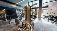 Located at 14 E Camden St in Hackensack, NJ, this well-appointed fitness center offers a sophisticated and fully equipped workout environment designed to meet a wide range of training needs. The space is organized into distinct exercise zones, each thoughtfully designed with both function and aesthetics in mind.

The cardio zone features a row of modern treadmills and elliptical cross-trainers positioned against a striking exposed brick wall, illuminated by sleek diagonal linear lighting that adds a dynamic visual element. Adjacent to this area, a selection of air bikes and additional cardio machines are bathed in natural light streaming through expansive floor-to-ceiling windows that overlook the surrounding urban streetscape.

The strength and functional training area is equipped with professional-grade cable and multi-station machines, along with a full dumbbell rack reflected in large wall-mounted mirrors. Colorful medicine balls and functional training tools add a vibrant touch to the space.

The overall design blends industrial and contemporary styles, combining exposed brick, textured concrete columns, patterned carpet tiles, and black-framed glass partitions. A mezzanine-level overhang and curated artwork on the walls contribute to the elevated ambiance, making this fitness center as visually engaging as it is practical for daily training.