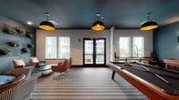 This well-appointed game and social lounge is designed for both recreation and relaxed gathering. The space is anchored by a full-size pool table with a dark felt surface and warm wood frame, positioned beneath a pair of striking oversized pendant lights with bold black-and-gold interiors that draw the eye upward to the deep teal-painted ceiling—a dramatic design choice that sets a sophisticated, energetic tone throughout.

Along one wall, a wet bar area features white subway tile backsplash, open shelving with decorative accents, a sink, and under-counter refrigeration, making the space well-suited for social events. Adjacent to the bar, a card or game table surrounded by houndstooth-patterned chairs provides a dedicated spot for tabletop gaming. A shuffleboard table is also visible, further enriching the recreational offerings.

A cozy lounge nook features woven rattan swivel chairs with teal cushions gathered around a low table, with a living wall of potted plants mounted on a dark pegboard panel adding a natural, textural element. Striped carpeting runs throughout, softening the space and tying the design together. Large windows and glass doors flood the room with natural light and offer views of the surrounding exterior, while a separate glass-enclosed room provides an additional private area within the lounge.