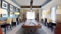 This single-story amenity space showcases a thoughtfully designed interior that blends contemporary and industrial aesthetics. At the heart of the space is a sweeping curved bar counter finished with light granite and dark cabinetry, lined with rustic wooden bar stools on metal frames. A cluster of glass pendant lights hangs overhead, adding a warm, layered glow to the area. Recessed lighting complements the pendants throughout, illuminating the light-toned tile flooring that runs seamlessly across the space.

Beyond the bar, the layout opens into a lounge and game room area anchored by a substantial wooden pool table, positioned beneath a striking overhead light fixture with exposed Edison bulbs. A textured slate-toned accent wall adds depth and contrast to the neutral palette, while exposed wood beams on the tray ceiling introduce a rustic element. Open metal shelving units display curated décor, and framed black-and-white artwork adorns the walls.

Seating areas with high-top tables and upholstered chairs are positioned throughout, creating versatile gathering spots. Sheer curtain-draped windows allow natural light to filter in, enhancing the welcoming ambiance. Overall, the space is designed for social interaction and leisure, offering a polished yet relaxed environment.