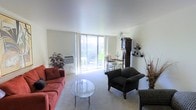 This single-floor residence centers around a generously sized living area filled with natural light streaming through a large sliding glass door that opens to an outdoor balcony or patio, offering views of lush greenery beyond. The neutral-toned carpet and cream-painted walls create a clean, airy backdrop throughout the space. A ceiling fan is positioned overhead, contributing to both function and a relaxed ambiance.

The living area is thoughtfully arranged with a terracotta-red upholstered sofa, a patterned armchair, and a circular glass-top coffee table, lending the room a casual yet classic character. A stately grandfather clock stands prominently along one wall, serving as a distinctive architectural accent and focal point. Adjacent shelving displays decorative items, adding a personalized touch to the space.

A dining area is visible beyond the living room, positioned near the sliding glass doors to take advantage of the natural light. The wall-mounted heating unit beneath the window ensures year-round comfort. Decorative plants placed throughout the living area add warmth and a natural element to the overall design.