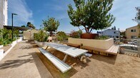 Located at 4340 44th Street in San Diego's City Heights neighborhood, this outdoor community space serves as a vibrant garden corridor nestled alongside a modern multi-story residential building. The space is thoughtfully designed to encourage community gathering and urban agriculture, stretching as a long, well-organized outdoor passageway filled with greenery and gathering amenities.

Raised garden beds constructed from whitewashed concrete block line the pathway and overflow with a diverse array of plants, including fruit trees, grapevines, ornamental grasses, herbs, and vegetables. Large galvanized metal containers and additional planter boxes punctuate the space, offering further growing areas and visual interest. Mature trees provide natural shade and contribute to a lush, park-like atmosphere under the sunny San Diego sky.

The hardscaped walkway is complemented by warm wood-toned composite benches integrated into the planter walls, offering casual seating throughout. A picnic table with bench seating provides a dedicated gathering spot, ideal for communal meals or outdoor activities. Street-level lighting fixtures ensure the space remains accessible and welcoming at all hours.

The adjacent building features a contemporary façade with stucco cladding, bold accent colors, and large windows, adding architectural character to the setting. Overall, this outdoor space combines functional urban gardening with comfortable communal amenities, creating an engaging, green retreat within the city.